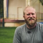 A cheerful bearded man smiling outdoors in a casual setting with greenery.